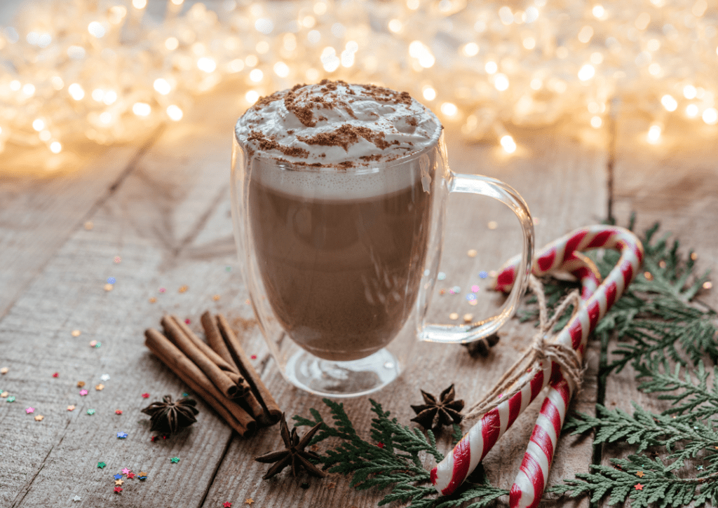 A festive mug of hot chocolate topped with whipped cream and a sprinkle of cinnamon, surrounded by holiday decorations and twinkling lights.