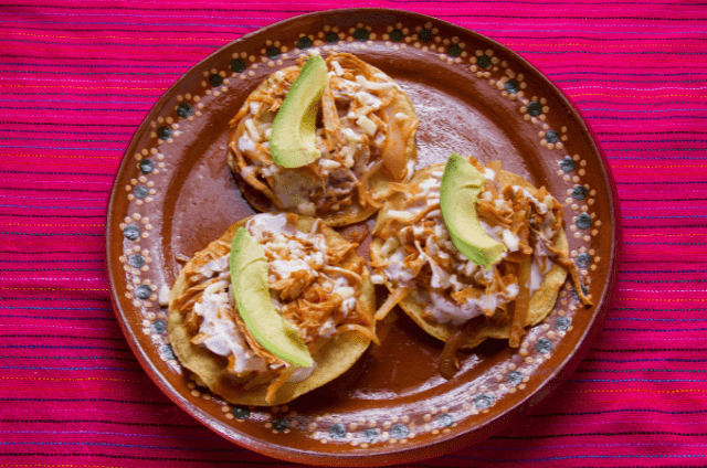 Tostadas topped with chicken tinga, avocado slices, and crumbled cheese on a traditional plate.