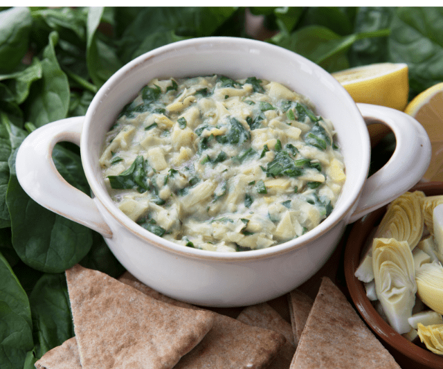 A creamy spinach and artichoke dip mixed with pasta served in a white bowl.