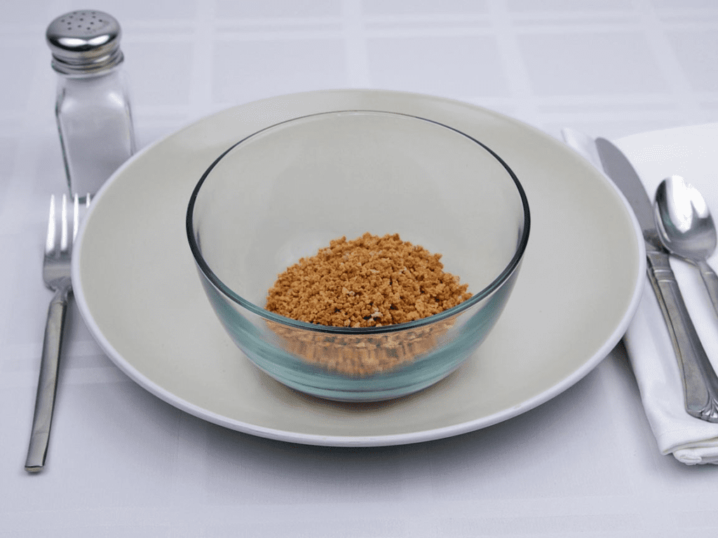 A small, clear bowl of brown, granulated cereal on a white plate with a fork and spoon on either side, and a salt shaker in the background.