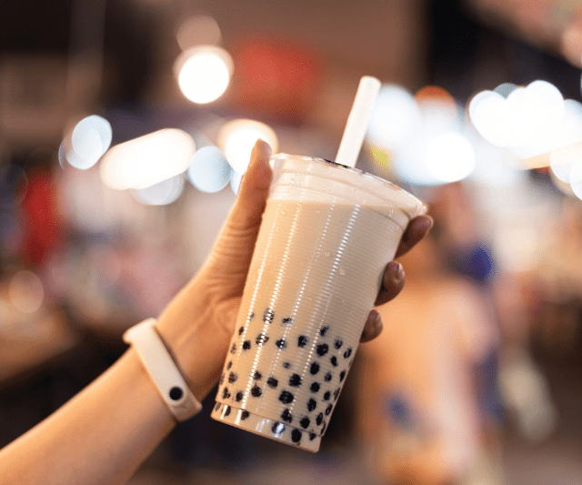A person's hand holding a plastic cup of bubble tea with a straw, with tapioca pearls visible at the bottom of the cup.