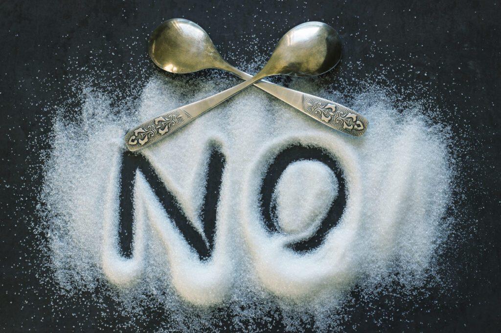 The word NO written into a pile of white granulated sugar with a black background. Two crossed spoons.