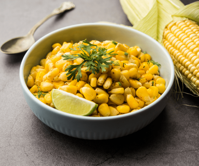 A bowl of cooked yellow corn kernels garnished with parsley and a lime wedge, with a whole corn cob and a spoon on the side. (Is Corn Good for Weight Loss?)