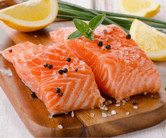 Two raw salmon fillets seasoned with black peppercorns and sea salt on a wooden cutting board, garnished with a sprig of basil and lemon wedges in the background. (Is Salmon Good for Weight Loss?)