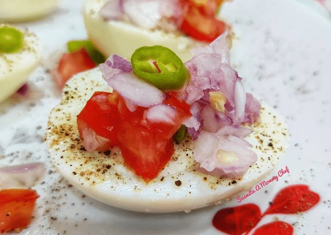A boiled egg halve topped with diced tomatoes, onions, and a slice of green chili, sprinkled with black pepper on a white plate with heart patterns.