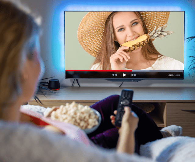 A woman watching a TV screen showing another woman eating a slice of pineapple.