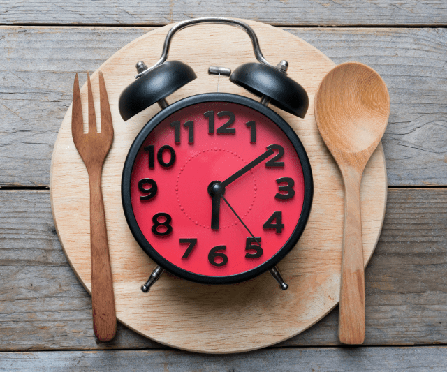 A red alarm clock set to 7 o'clock is placed on a wooden cutting board along with a wooden fork and spoon, symbolizing the concept of meal timing.