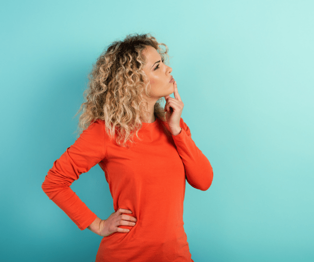 A woman with curly hair, wearing an orange long-sleeve shirt, standing with one hand on her hip and the other on her chin in a pondering gesture, against a turquoise background.