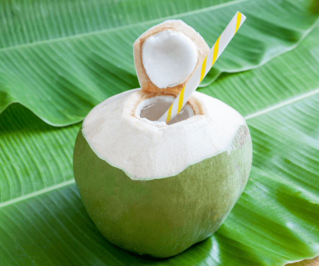 A fresh green coconut with a hole cut on top and a striped straw inserted, placed on a green leaf.
