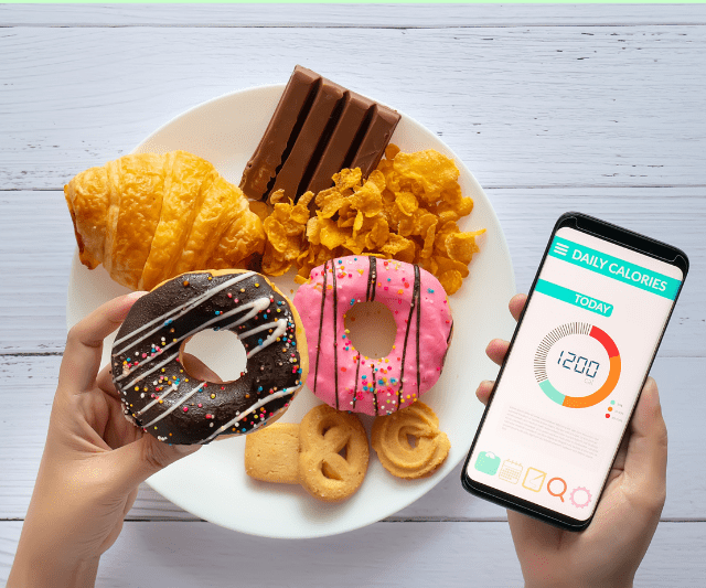 A person is holding a donut in one hand and a smartphone displaying a daily calorie tracking app in the other hand. The plate in front of them is filled with various high-calorie foods such as croissants, chocolate bars, cornflakes, and cookies.