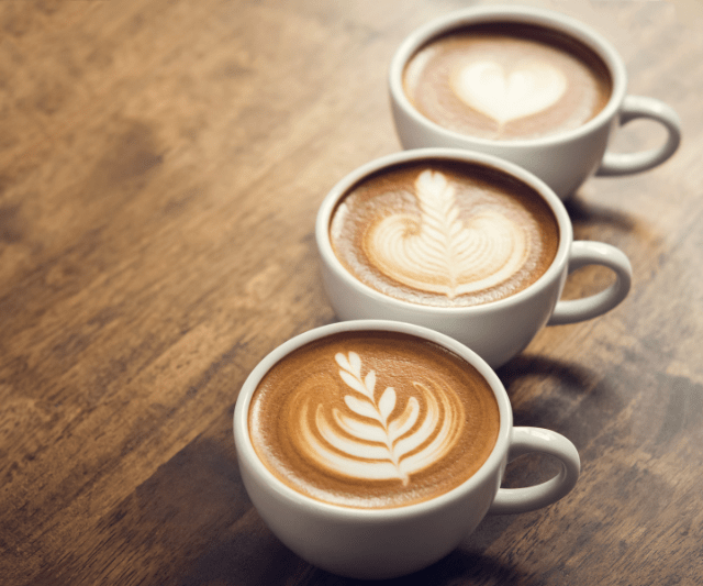 Three cups of coffee on a wooden table, each with intricate latte art designs on the surface, including a heart and two variations of leaf patterns.