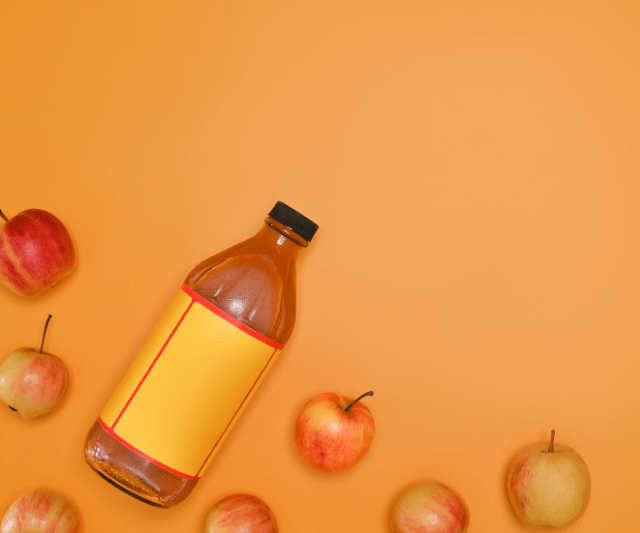 A bottle of apple cider vinegar with a blank yellow label, surrounded by several whole apples on an orange background.