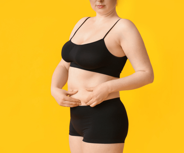 A woman in black sportswear pinching her belly fat, standing against a bright yellow background.