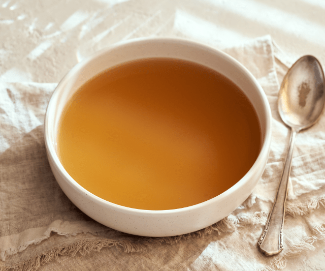 A bowl of clear bone broth with a spoon placed beside it on a light-colored fabric napkin.