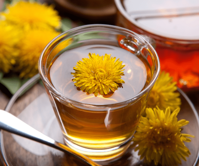 A close-up of a clear glass cup filled with dandelion tea, with a vibrant yellow dandelion flower floating on top. The background includes additional dandelion flowers and another cup of tea, creating a warm and inviting scene.