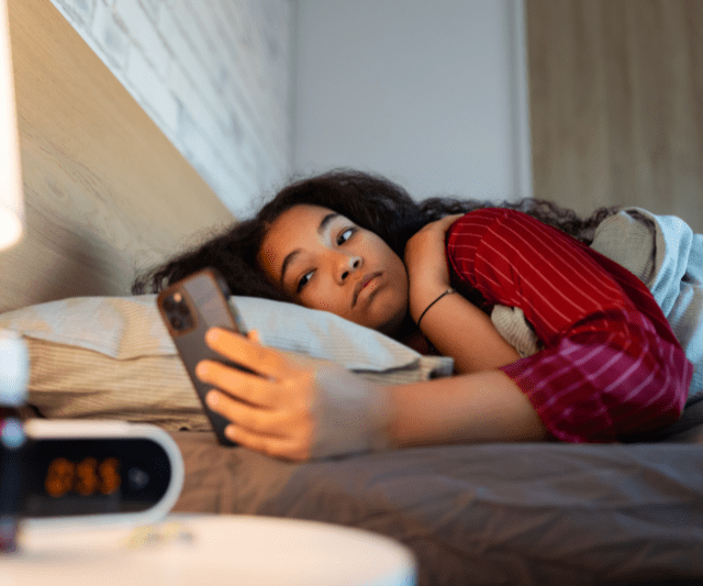 Woman lying in bed, looking at her phone, appearing tired.