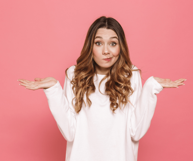 A young woman with long, wavy hair, wearing a white sweatshirt, stands against a pink background. She has a puzzled expression on her face, with her eyebrows raised and lips pursed, while shrugging her shoulders and holding her hands out to the sides.