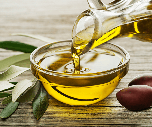 Olive oil being poured from a glass bottle into a small bowl, with olive branches and olives on a wooden surface.