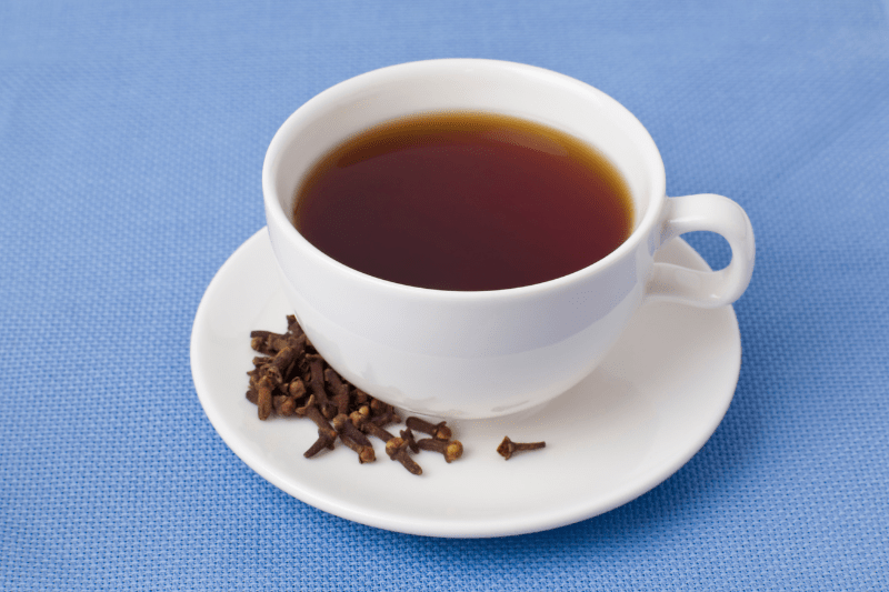 A cup of black tea with cloves on a white saucer placed on a blue tablecloth.