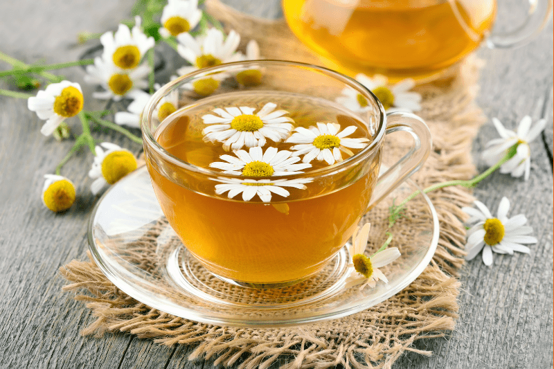 A clear glass cup of chamomile tea garnished with fresh chamomile flowers, placed on a rustic woven mat with more chamomile flowers scattered around.