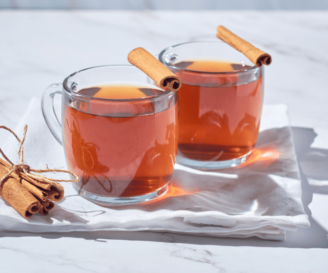 Two clear glass mugs filled with cinnamon tea, each garnished with a cinnamon stick, placed on a white cloth napkin with a small bundle of cinnamon sticks beside them.