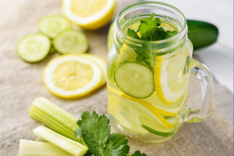 A mason jar filled with detox water containing lemon slices, cucumber slices, and fresh mint leaves. Surrounding the jar are additional lemon and cucumber slices, along with celery sticks and a parsley leaf. The setup is on a light-colored cloth.