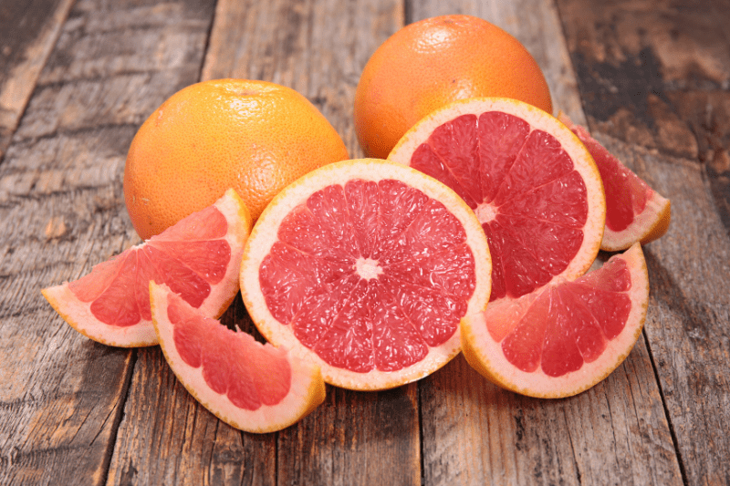 Several grapefruits on a wooden surface. Some are whole, while others are cut in half or into wedges, revealing their pink-red juicy flesh. The fruits have a vibrant orange-pink exterior and are arranged in a casual, appetizing display.