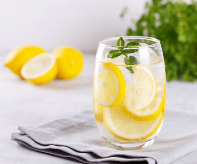 A glass of refreshing lemon water with slices of lemon and a sprig of mint, placed on a folded napkin, with whole lemons and green herbs in the background.