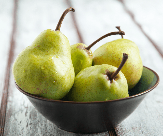 A brown bowl filled with fresh green pears, placed on a rustic white wooden surface.