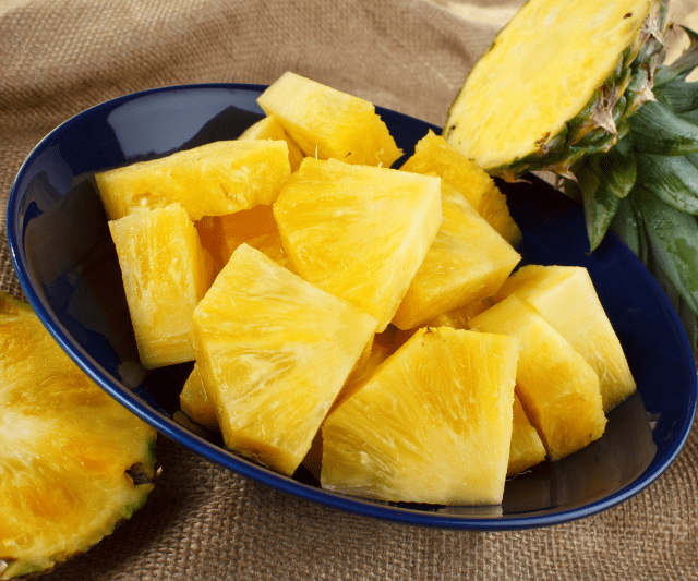 A bowl of fresh pineapple chunks, with a whole pineapple and pineapple slices in the background.